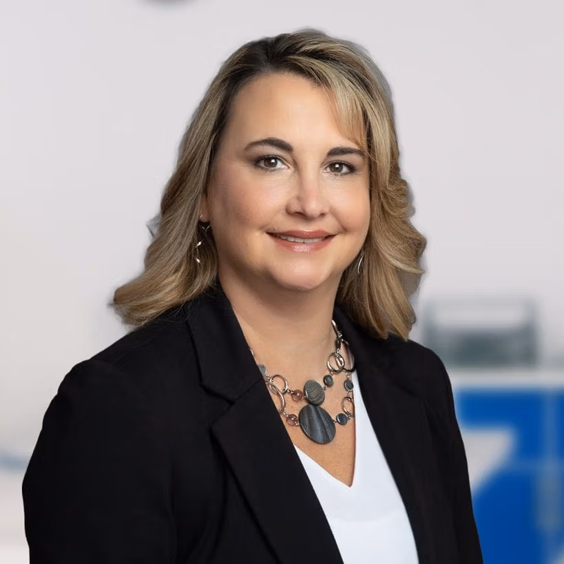 Amy Eveland, looking at the camera smiling with a black blazer on and muted blue background.