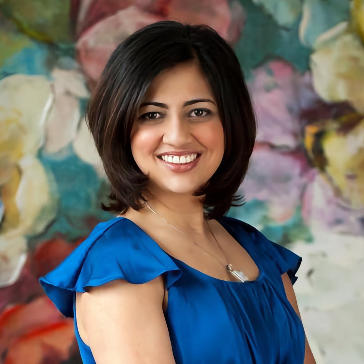 Sumbul Naqvi smiling in a blue dress, standing in front of a colorful backdrop.