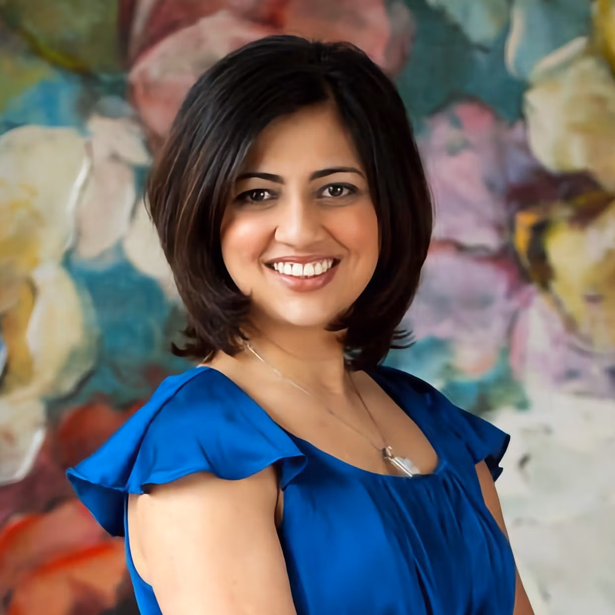 Sumbul Naqvi smiling in a blue dress, standing in front of a colorful backdrop.
